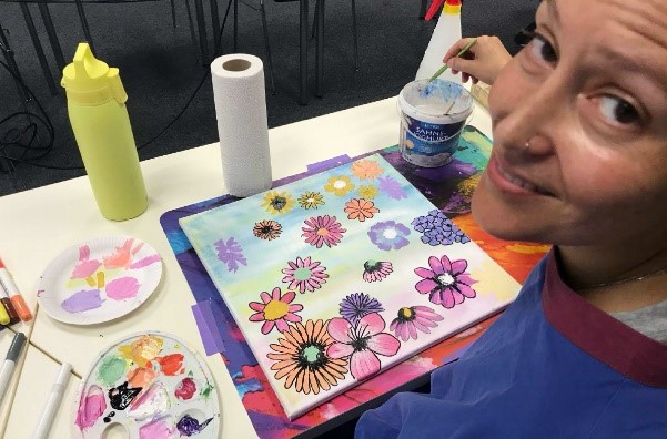 Farbraum_onkologisches_Kunstprojekt_Universitätsmedizin_Göttingen_Patientin_Blumenbild Auf dem Bild zu sehen ist eine Patienten im Rahmen des onkologischen Kunstprojektes Farbraum, die gerade ein farbenfrohes Bild mit vielen Blumen anfertigt und in die Kamera lächelt.