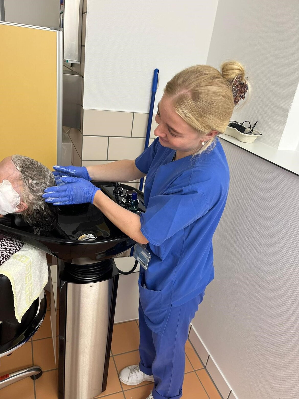 Auf dem Bild zu sehen ist eine Pflegerin, die einer Patientin am Haarwaschbecken die Haare wäscht.