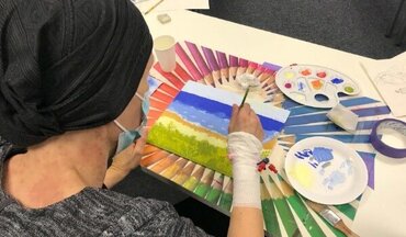 Farbraum_onkologisches_Kunstprojekt_Universitätsmedizin_Göttingen_Patientin_Landschaftsgemälde Auf dem Bild zu sehen ist eine Patienten im Rahmen des onkologischen Kunstprojektes Farbraum, die gerade ein Bild einer Landschaft anfertigt.